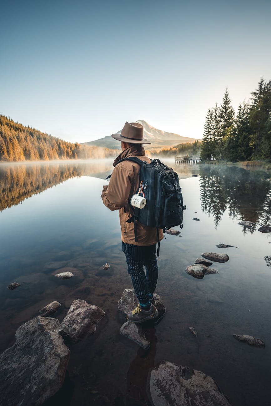 man in brown jacket and brown hat standing on rock near lake